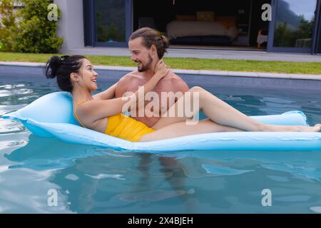 Birassische Frau und kaukasischer Mann, die sich draußen im Pool entspannen. Beide tragen eine Sonnenbrille, genießen den sonnigen Tag und lächeln einander an, unverfälscht Stockfoto