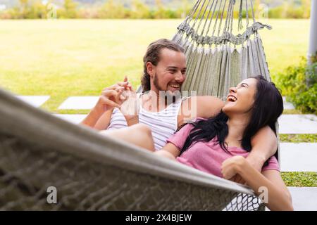 Verschiedene Paare lachen draußen in der Hängematte und genießen die gemeinsame Zeit. Zweirassige Frau mit langen schwarzen Haaren und kaukasischer Mann mit Bart, beide jung, rel Stockfoto