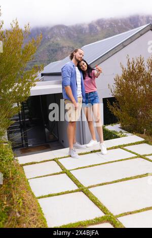 Vielfältige Paare, die sich im Freien tummeln und ihr neues Zuhause genießen. Zweirassige Frau mit langen dunklen Haaren und kaukasischer Mann mit Bart lächeln warm, unalt Stockfoto