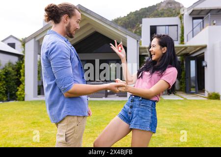 Zweirassige Frau und kaukasischer Mann, neue Hausbesitzer, lachen draußen. Hände haltend, sie mit langen schwarzen Haaren, er in blauem Hemd, beide sehen jung aus Stockfoto