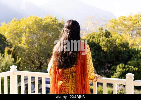 Indische junge Frau, draußen auf dem Balkon, die Natur anschauend. In einem traditionellen Outfit in leuchtendem Orange, mit langen schwarzen Haaren, die im Wind fließen, unverändert Stockfoto