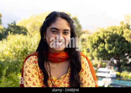 Eine junge indische Frau im gelben Outfit draußen, mit rotem Schal, lächelnd. Mit langen schwarzen Haaren und dunklen Augen genießt sie einen klaren Teint und einen Stockfoto