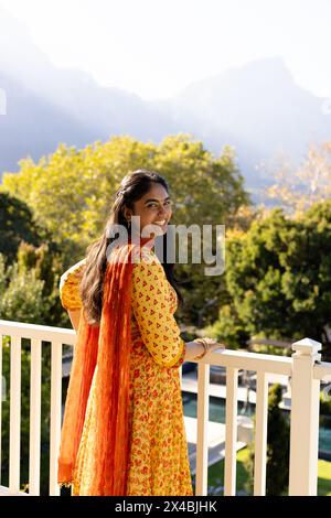 Indische Frau in gelbem Kleid, steht draußen auf dem Balkon und genießt den Blick auf die Berge. Mit langen schwarzen Haaren und warmem Lächeln, frische Luft und Ruhe Stockfoto