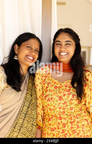 Indische Mutter und Teenager-Tochter, beide lächeln, stehen zuhause zusammen. Mutter zeigt graue Streifen in ihrem dunklen Haar und hält Hände mit ihr Stockfoto