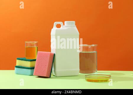 Vorderansicht des weißen Waschmittelkanisters mit bunten Schwämmen und Glaswaren gefüllt mit gelber Flüssigkeit auf Orangenhintergrund. Modellkanister Stockfoto