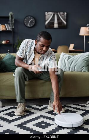 Vertikale Aufnahme des jungen Schwarzen Mannes, der auf dem Sofa sitzt und einen modernen Staubsauger anschaltet Stockfoto