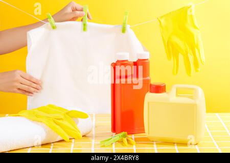 Auf gelbem Hintergrund trocknen die Hände der Frau ein frisch gewaschenes weißes Handtuch, Gummihandschuhe und Plastikkanister verziert. Mockup-Szene, Flaschenwitz Stockfoto
