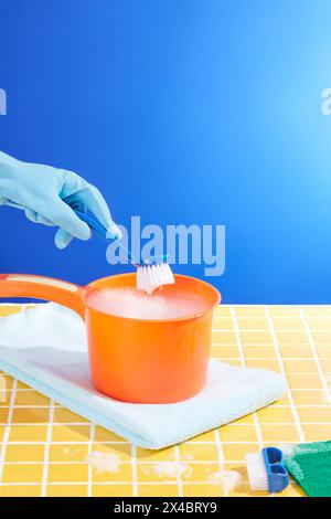 Vorderansicht des orangefarbenen Kunststoff-Wasserlöffelbehälters Reinigungsschaum und Wischers auf gelbem Keramikhintergrund. Hand mit Gummihandschuh hält Stockfoto