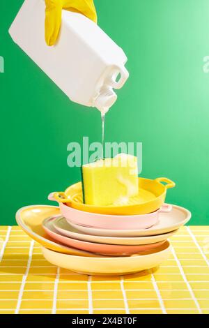 Stapel von Keramikgeschirr und Pfannen, die auf dem gelben Fliesenboden gewaschen werden. Eine Hand hält den Kunststoffbehälter und gießt das Reinigungsmittel langsam herunter. Profe Stockfoto