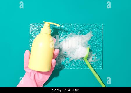 Eine Hand, die einen rosafarbenen Gummihandschuh trägt, hält eine gelbe Plastikflasche mit Sprühspitze, weißem Schaum und einer Toilettenbürste auf einer Glasplatte. Simulierte Scen Stockfoto