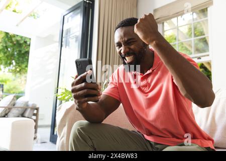 Afroamerikaner feiert, während er zu Hause auf das Smartphone schaut. Er trägt ein rosafarbenes Hemd, hat kurze schwarze Haare und einen Bart, unverändert. Stockfoto