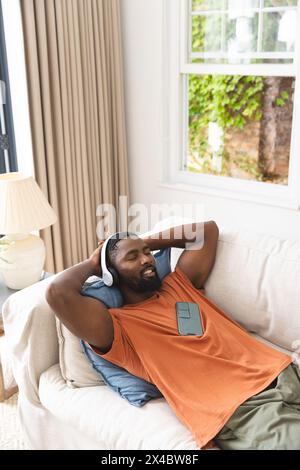 Afroamerikanischer Mann, der sich auf der Couch entspannt, Kopfhörer trägt, Kopierraum. Lässige Kleidung tragen, Smartphone halten, kurze schwarze Haare, blauer Hintergrund Stockfoto