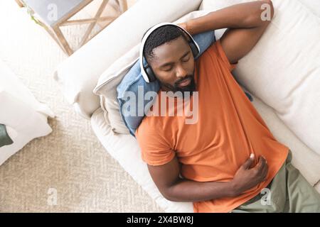 Afroamerikanischer männlicher Hausbesitzer, der sich auf der Couch entspannt, Musik genießt, Kopierraum. Er trägt ein orangefarbenes Hemd, Kopfhörer und hat unveränderten Bart Stockfoto