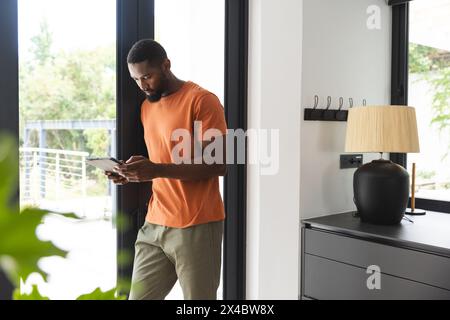 Ein junger Afroamerikaner, der eine Tafel in der Nähe des Fensters hält. In lässigen Klamotten konzentriert er sich auf den Bildschirm, steht drinnen, unverfälscht Stockfoto