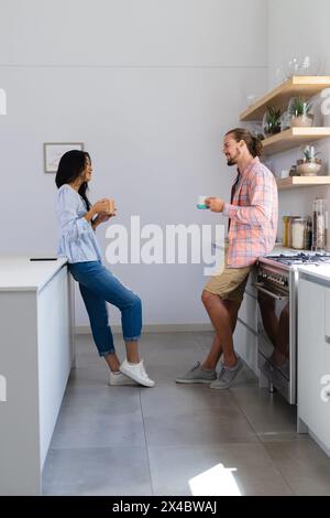 Vielfältiges Paar, das Kaffee in der modernen Küche zu Hause genießt. Sie hat lange schwarze Haare, er trägt einen Bart und lange Haare, unverändert Stockfoto