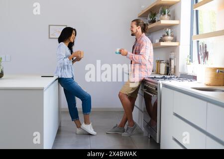 Birassische Frau und kaukasischer Mann, Paar, genießen Kaffee in der Küche zu Hause. Beide haben dunkles Haar, legere Kleidung und lächeln einander an, unal Stockfoto