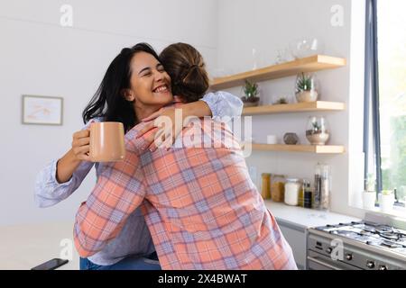 Diverse Paare, die sich zu Hause in der Küche umarmen, eine Frau, die einen Becher hält. Birassische Frau hat lange schwarze Haare, kaukasischer Mann trägt kariertes Hemd, unverändert Stockfoto