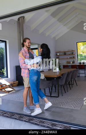 Ein vielseitiges Paar, das sich zu Hause umgibt und beide ein warmes Lächeln zeigt. Sie teilt sich einen gemütlichen Moment vor weißem Hintergrund, sie mit langen schwarzen Haaren, er mit einem Bären Stockfoto