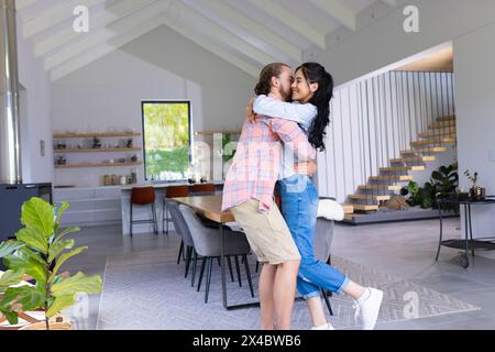 Vielseitiges Paar, das sich in einem modernen Zuhause umgibt. Zweirassige Frau mit langen dunklen Haaren und kaukasischer Mann mit Bart, die einen liebenden Moment teilen, unverfälscht Stockfoto
