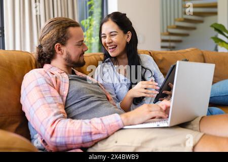 Zweirassige Frau und kaukasischer Mann, Paar, lachend und auf Laptop zu Hause. Sie hat dunkle Haare und er trägt Bart und langes Haar, beide gekleidet ca. Stockfoto
