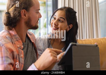 Ein vielseitiges Paar, das sich ein Tablet anschaut und zu Hause auf der Couch sitzt. Zweirassige Frau mit langen schwarzen Haaren, kaukasischer Mann mit lockigen Haaren, unverändert Stockfoto