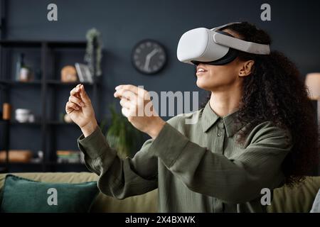 Mittlere Nahaufnahme einer jungen Frau, die ein VR-Headset trägt und im Wohnzimmer auf dem Sofa sitzt, Videospiele spielt oder arbeitet Stockfoto