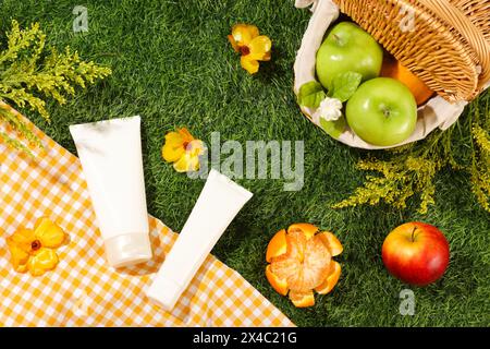 Blick von oben auf Bambuskorb voll mit Bio-Früchten auf grünem Grashintergrund mit kariertem Stoff und gelben Blumen. Zwei nicht mit Mo gekennzeichnete Kosmetikröhrchen Stockfoto