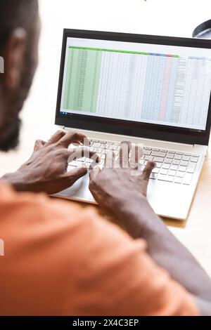 Zu Hause schreibt ein männlicher afroamerikanischer Hausbesitzer auf einem Laptop und sieht sich eine Tabelle an. Tragen Sie ein orangefarbenes Hemd und konzentrieren Sie sich intensiv auf die Arbeit mit einem orangefarbenen B Stockfoto