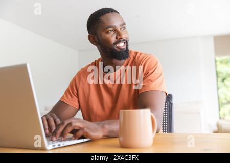 Zu Hause, ein männlicher Afroamerikaner, der einen Laptop benutzt und wegblickt. Ein lässiges orangefarbenes Hemd, sanft lächelnd, mit kurzem Bart, gegen einen Orang Stockfoto