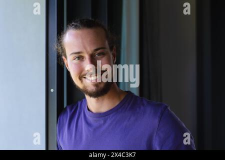 Ein junger kaukasischer Mann, der sich zu Hause an den Türrahmen lehnt. Mit einem Bart, langen braunen Haaren und blauen Augen strahlt er eine entspannte Atmosphäre aus, unalt Stockfoto
