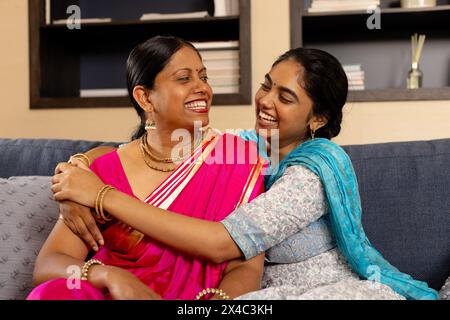Indische Mutter und Tochter im Teenageralter sitzen zusammen und lachen zusammen. Beide mit dunklem Haar, Mutter in rosa Sari, Teenager-Tochter in blau, teilt Freude Stockfoto