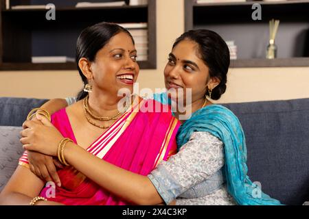 Indische Mutter und Tochter im Teenageralter, die sich in einem gemütlichen Zimmer lachen. Beide mit dunklem Haar, Mutter in hellem Sari, Tochter in Blau, genießt wieder Wärme Stockfoto
