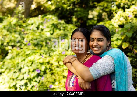 Indische Mutter und Teenager-Tochter umarmen, lächeln im Garten, Kopierraum. Beide haben dunkles Haar, mit Mutter kurz geschnitten und Tochter lang, unalt Stockfoto