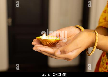 Indische junge Frau, die zu Hause eine beleuchtete Lehmlampe hält. Gelbes Kleid mit Mustern und goldenen Armreifen, dunkles Haar, Teenager-Tochter feiert Stockfoto