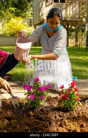 Indische Mutter und Teenager-Tochter im Garten, tragen rote Kurta, graben Erde. Teenager-Tochter tränkt Blumen, genießt den sonnigen Tag mit rotem Backgrou Stockfoto