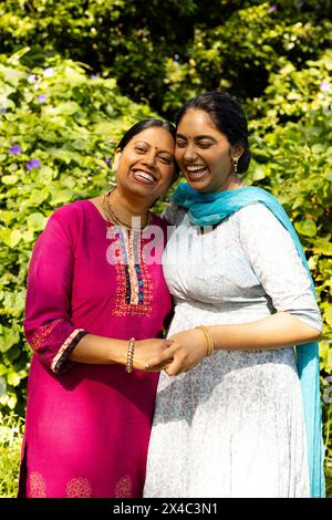 Indische Mutter und Teenager-Tochter lachen zusammen im Garten. Beide tragen traditionelle Kleider, Mutter in rosa Hintergrund und Teenager-Tochter in bl Stockfoto
