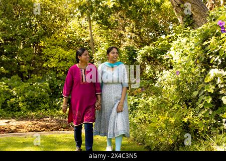 Indische Mutter und Teenager-Tochter gehen im Garten, beide sehen glücklich aus. Mutter mit schwarzen Haaren, die einen Hauch von Grau zeigt, und Tochter im Teenageralter mit Lon Stockfoto