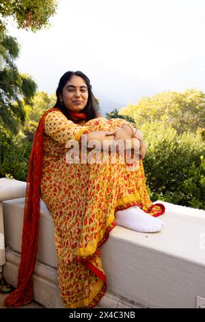 Die junge indische Frau sitzt draußen und trägt gelben Sari. Sie hat langes schwarzes Haar, lächelt und sieht etwas aus, das unverfälscht ist Stockfoto