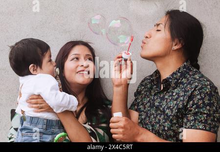 Eine ecuadorianische Familie mit einem Vater mit einem traditionellen Flechtflecht genießt einen verspielten Moment mit Seifenblasen und ihrem Baby. Stockfoto