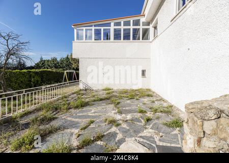 Eine raue weiße Zementwand mit verglastem Erkerfenster die Seitenfassade eines Einfamilienhauses mit ungepflegtem Unkraut Stockfoto