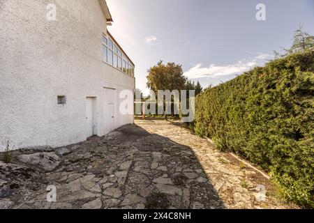 Ein Steinweg an der Seite eines Einfamilienhauses mit weißer Zementfassade und umlaufendem Heckenzaun Stockfoto