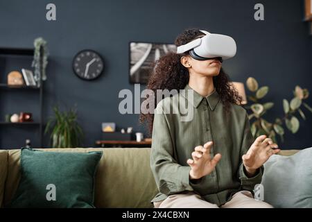 Mittlere Aufnahme einer jungen Frau mit VR-Headset, die im Wohnzimmer auf der Couch sitzt und online arbeitet Stockfoto