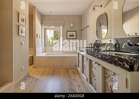 Schönes und geräumiges Badezimmer mit schwarzer Marmorplatte mit weißen Adern, zwei Spiegeln, großer Badewanne unter einem Fenster und separater Dusche und Toilette Stockfoto