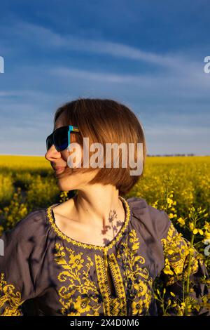 Glückliche Frau, 35-40 Jahre alt, in blauer Sonnenbrille und einer ukrainischen bestickten Bluse auf einem Rapsfeld. Digitale Detox, Sommerwochenende, genießen Sie den Moment Stockfoto