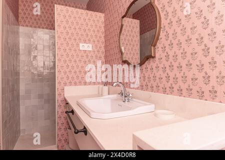 Bad mit cremefarbener Marmorplatte, goldgerahmten Spiegeln, rosafarbener Tapete, heller, ebenerdiger Dusche und weißem Porzellanwaschbecken Stockfoto