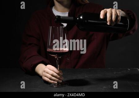 Alkoholsucht. Frau, die Rotwein aus der Flasche ins Glas gießt, an einem dunklen, strukturierten Tisch, Nahaufnahme Stockfoto