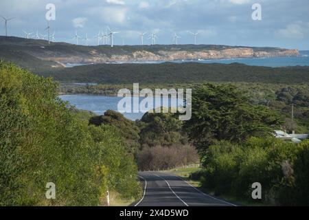 Landschaft mit einer Straße im Vordergrund und Windturbinen Farm an einer Seeküste vor dem Hintergrund von grünen Hügeln und bewölktem Himmel Stockfoto