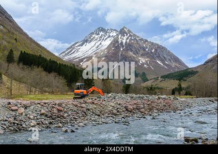 Ein Bagger arbeitet an einem Flussufer mit einem schneebedeckten Berg im Hintergrund. Stockfoto