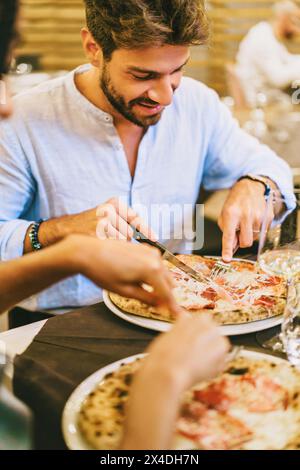 Junger Mann - Schneiden und Genießen von Pizza in einem pulsierenden Restaurant, zwangloses Abendessen mit Freunden - Genuss, gesellig. Stockfoto