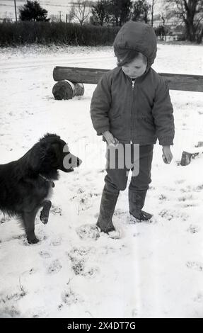 1960er Jahre, historisch, Winterzeit und ein kleiner Junge spielt mit seinem Hund draußen im Schnee. Stockfoto